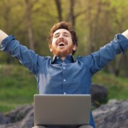 Ein Mann mit Laptop auf - Beruflicher Wechsel dem Schoß im Wald wirft glücklich die Arme hoch und freut sich über neuen Job