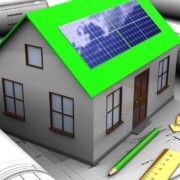 Ein Modell eines Hauses mit grünem Dach und Photovoltaikanlage steht auf einem Bauplan, rundherum liegen weitere Rollen Baupläne, Stifte und Lineale | Energieeffizient bauen