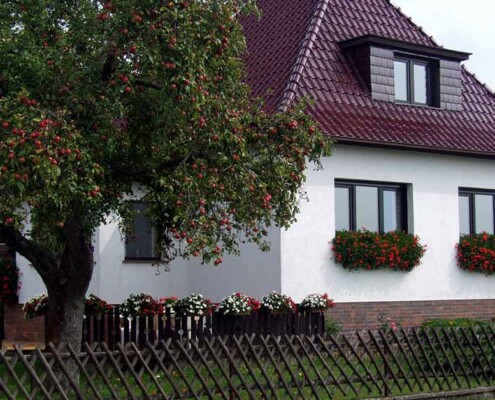 Ein Haus mit weißer Fassade und rotem Dach, mit einem Baum im Vorgarten und einem Holzzaun zur Straße | Fehler Immobilienverkauf