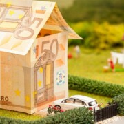 Modellhaus aus 50 Euro Scheinen in einem Garten mit einem weißen Spielzeugauto vor der Tür | Immobilienfinanzierung