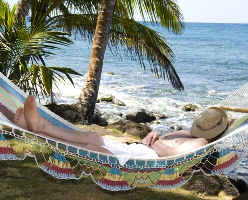 Ein Mann liegt entspannt in seiner Hängematte am Strand im Ausland unter Palmen, mit Hut auf dem Gesicht, und genießt sein Leben, weil er weiß, dass ein Qualitätsmakler sich um den Verkauf seiner Immobilie in Deutschland kümmert | Immobilienverkauf