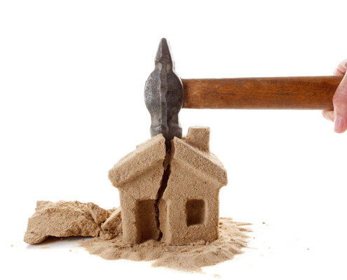 Eine Hand schlägt mit einem Hammer auf ein Haus aus Sand und zerstört es | Notverkauf Immobilie