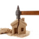 Eine Hand schlägt mit einem Hammer auf ein Haus aus Sand und zerstört es | Notverkauf Immobilie