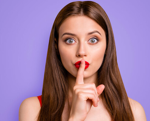 ein junge Frau, die sich den Zeigefinger vor den Mund hält um Verschwiegenheit zu demonstrieren