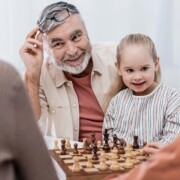 Wohnen im Alter Ein Seniorenpärchen sitzt mit seinen Enkelkindern glücklich im Wohnzimmer und spielt Schach als Symbolbild für strategisch kluge Entscheidungen bei Investments für die Altersvorsorge | Wohnen im Alter