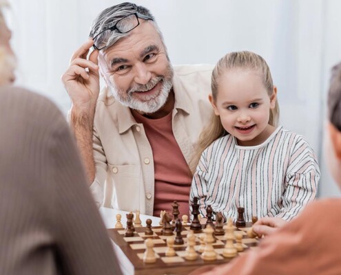 Ein Seniorenpärchen sitzt mit seinen Enkelkindern glücklich im Wohnzimmer und spielt Schach als Symbolbild für strategisch kluge Entscheidungen bei Investments für die Altersvorsorge | Wohnen im Alter