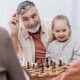 Ein Seniorenpärchen sitzt mit seinen Enkelkindern glücklich im Wohnzimmer und spielt Schach als Symbolbild für strategisch kluge Entscheidungen bei Investments für die Altersvorsorge | Wohnen im Alter