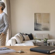 Ältere Frau steht an einem großen Fenster in einem hellen Wohnzimmer; auf dem Tisch im Vordergrund liegen Immobilienunterlagen, ein Schlüsselbund und ein Ordner - als Symbolbild für einen Immobilienverkauf mit eingetragenem Wohnrecht | Immobilienverkauf