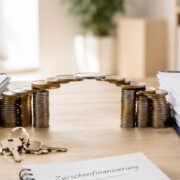 Münzstapel in Form eines Hauses zwischen Immobilienunterlagen und Schlüsseln symbolisieren eine Zwischenfinanzierung beim Immobilienkauf. I Zwischenfinanzierung